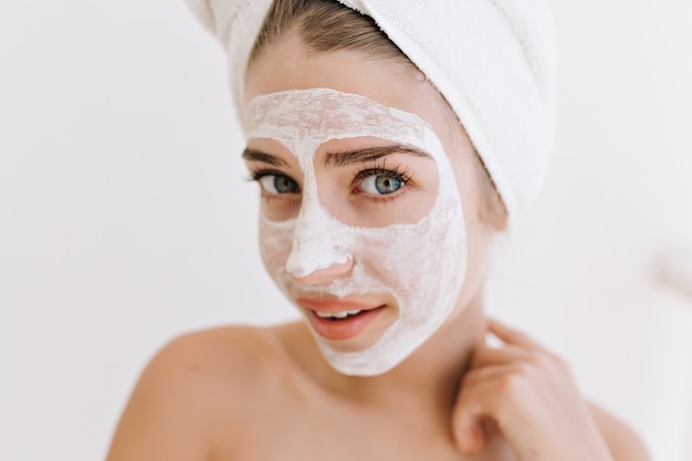 Il ritratto del primo piano di giovane e bella donna con gli asciugamani dopo il bagno fa la maschera cosmetica sul viso.