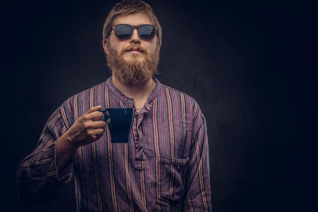 Il ritratto del primo piano di un ragazzo hipster che indossa occhiali da sole vestito con una camicia vecchio stile tiene una tazza di caffè. Isolato su uno sfondo scuro.