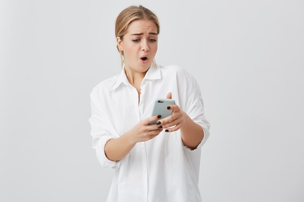 Il ritratto del primo piano il giovane caucasico femminile ansioso o colpito con capelli biondi che esaminano il telefono che vede le cattive notizie o le foto con emozione disgustosa sul suo fronte ha isolato il fondo grigio.