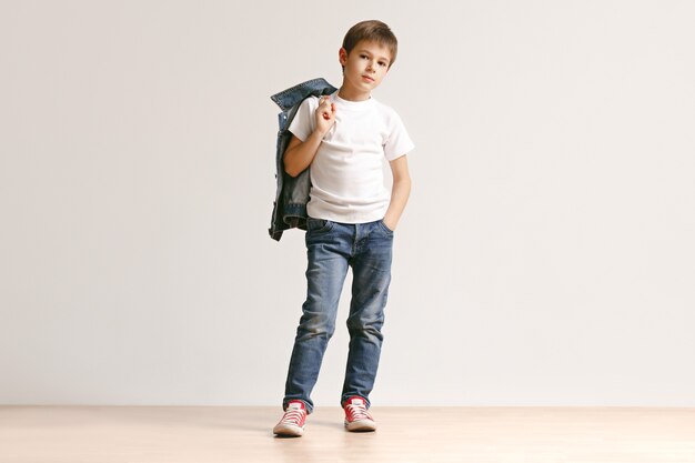 Il ritratto del ragazzino sveglio in vestiti di jeans alla moda che guarda l'obbiettivo in studio