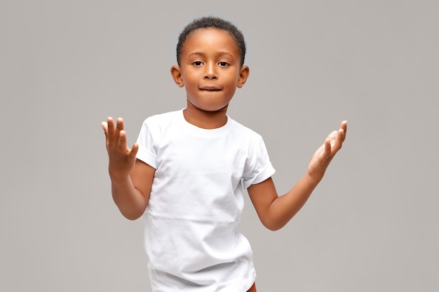 Il ritratto del ragazzo afroamericano sveglio freddo si è vestito in maglietta bianca casuale che ha espressione facciale sicura che mostra qualche gesto con le mani, labbro inferiore mordace. Bambini e concetto di stile di vita