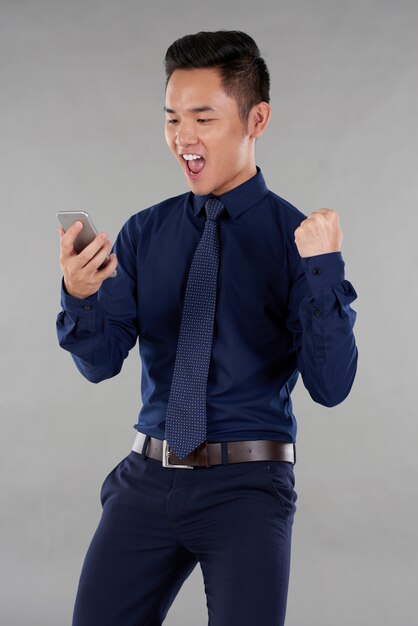 Il ritratto del tipo asiatico in vestiti astuti ha eccitato dalle notizie dello smartphone contro fondo grigio