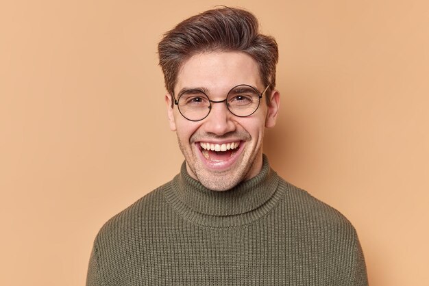 Il ritratto dell'uomo adulto bello felice sorride con gioia guarda direttamente la telecamera ridacchia positivamente ha l'acconciatura indossa occhiali rotondi maglione casual isolato su sfondo beige. Concetto di emozioni