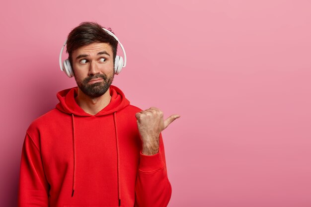 Il ritratto dell'uomo barbuto curioso punta il pollice nello spazio vuoto a destra, indossa l'auricolare stereo e una felpa casual rossa, dimostra qualcosa di interessante, isolato sul muro rosa pastello.