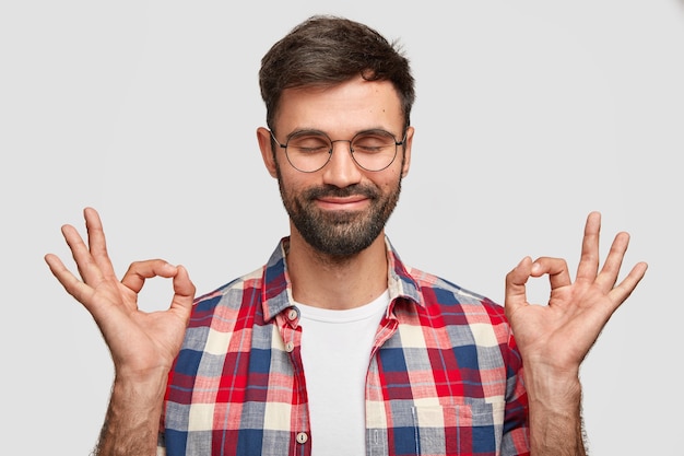 Il ritratto dell'uomo bello con la barba lunga mostra bene o come gesto con entrambe le mani, sorride piacevolmente, tiene gli occhi chiusi