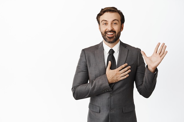 Il ritratto dell'uomo corporativo che alza il palmo ha messo la mano sul cuore e sorridendo sinceramente facendo la promessa si presenta in piedi in abito grigio su sfondo bianco