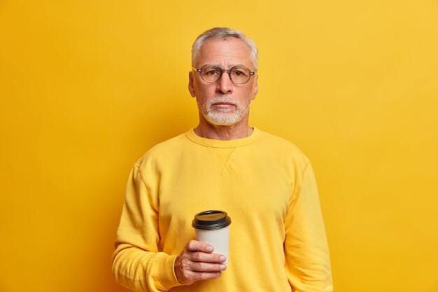 Il ritratto dell'uomo dai capelli grigi barbuto tiene la tazza di caffè da asporto vestita in ponticello casuale guarda direttamente nella parte anteriore isolata sopra la parete gialla soddisfatta con il tempo libero