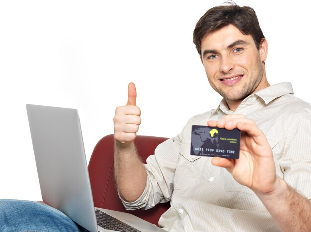 Il ritratto dell'uomo felice sorridente con il computer portatile dà i pollici in su e mostra la carta di credito isolata su bianco.