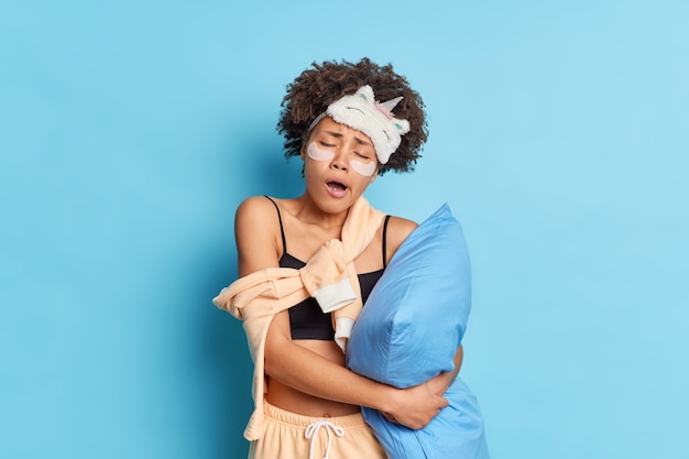 Il ritratto della donna afroamericana dai capelli ricci sonnolenta sbadiglia dopo il risveglio precoce tiene il cuscino vestito in pigiama e la maschera del sonno inclina la testa applica pastiglie di collagene sotto gli occhi isolati sopra il muro blu