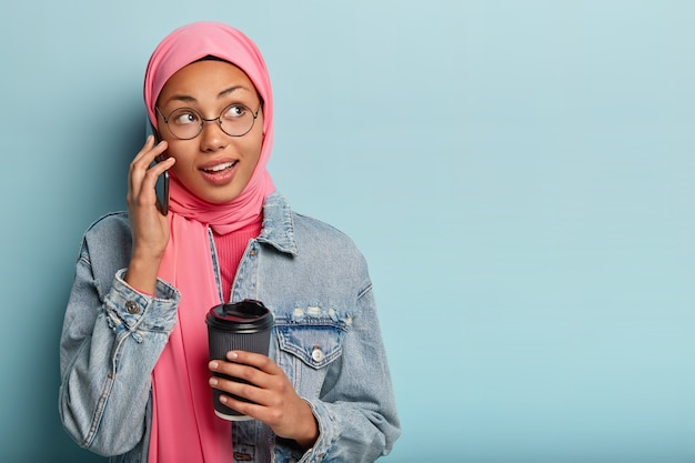 Il ritratto della donna araba felice fa la disposizione tramite il telefono cellulare, tiene la tazza di caffè di carta, riceve la buona offerta, guarda da parte