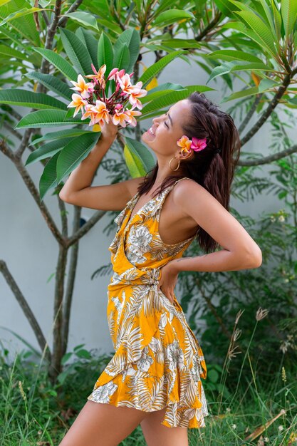 Il ritratto della donna asiatica in vestito giallo da estate sta con il fiore tailandese di plumeria nei capelli e negli orecchini rotondi