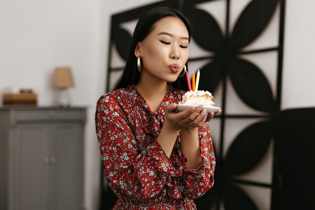 Il ritratto della donna castana in vestito floreale rosso spegne le candele alla torta di compleanno cremosa cream