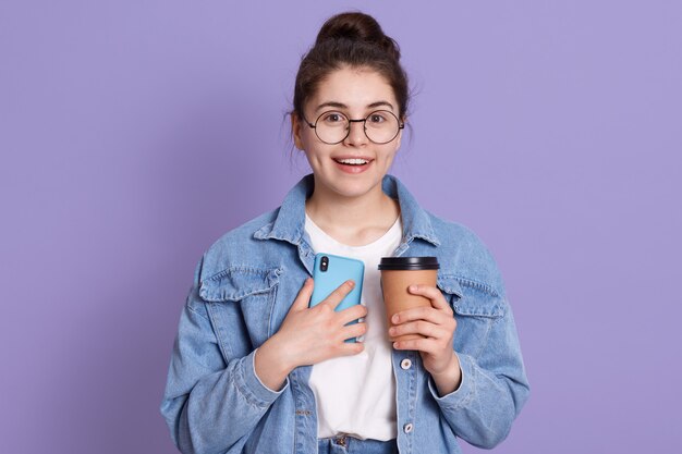 Il ritratto della donna caucasica sorridente che sta contro la parete lilla con porta via il caffè