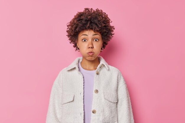 Il ritratto della donna dai capelli ricci sorpresa soffia le guance trattiene il respiro dallo stupore vestito con una pelliccia bianca isolata sopra la parete rosa