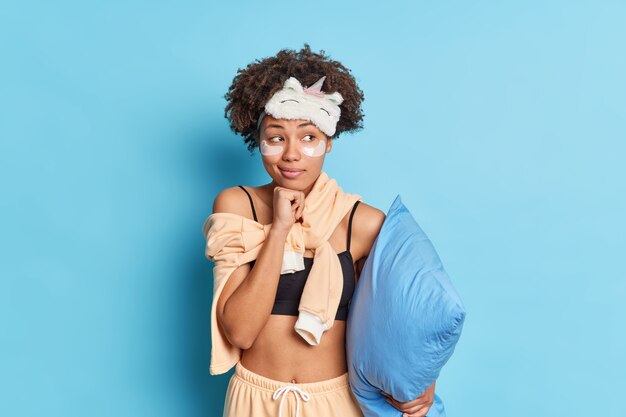 Il ritratto della donna dai capelli ricci tiene la mano sotto il mento guarda pensieroso da parte vestito in indumenti da notte tiene il cuscino andando ad avere sonno isolato sopra la parete blu