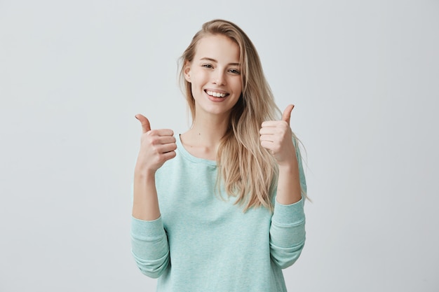 Il ritratto della donna femminile bionda con il vasto sorriso ed i pollici aumentano il gesto