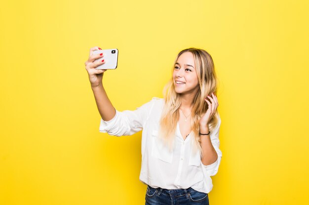 Il ritratto della donna prende selfie che tiene il telefono astuto in mano che spara selfie isolato sulla parete gialla