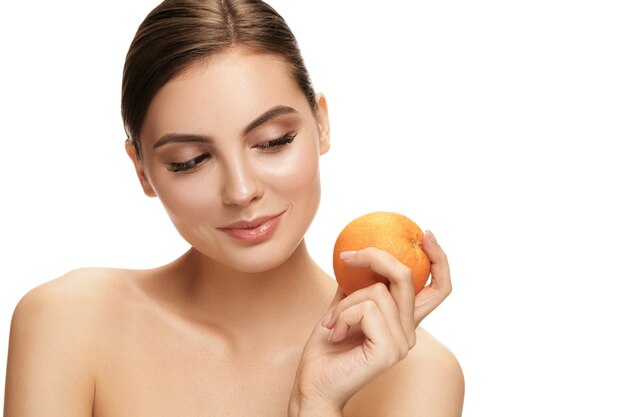 Il ritratto della donna sorridente caucasica attraente isolata sul muro bianco con frutta arancione.