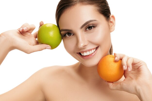 Il ritratto della donna sorridente caucasica attraente isolata sulla parete bianca dello studio con la mela verde e la frutta arancione. La bellezza, la cura, la pelle, il trattamento, la salute, la spa, i cosmetici e il concetto di annuncio