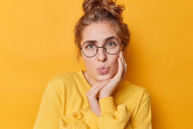 Il ritratto della giovane donna adorabile e tenera ha le labbra piegate tiene la mano sulla guancia guarda delicatamente la macchina fotografica indossa occhiali rotondi trasparenti e il maglione posa su sfondo giallo ha un'espressione romantica