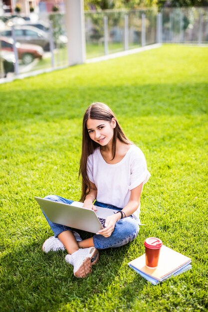 Il ritratto della giovane donna attraente che si siede sull'erba verde in parco con le gambe ha attraversato durante il giorno di estate mentre per mezzo del computer portatile