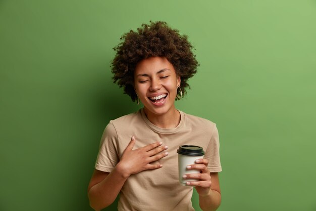 Il ritratto della ragazza etnica allegra si diverte e ride ad alta voce, tiene la tazza di caffè usa e getta, sorride ampiamente e chiude gli occhi, ha una conversazione allegra, isolata sul muro verde. Concetto di stile di vita