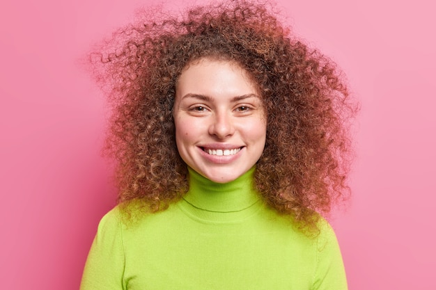 Il ritratto di bella donna europea allegra con i capelli folti ricci sorride ampiamente vestito in dolcevita verde isolato sopra la parete rosa. La donna europea sorridente spensierata si gode il tempo libero