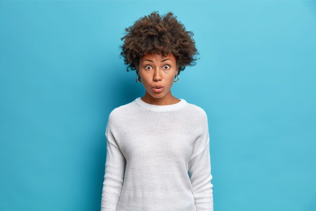 Il ritratto di bella donna scioccata confusa guarda sorprendentemente alla telecamera imitata di essere accusato o incolpato indossa un maglione bianco casual isolato sopra il muro blu