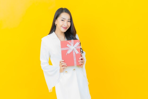 Il ritratto di bella giovane donna asiatica sorride con il contenitore di regalo rosso sulla parete gialla