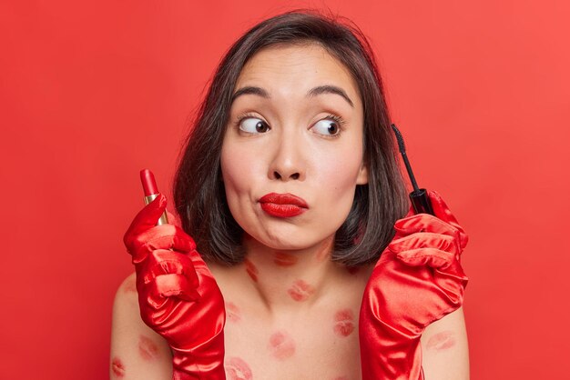 Il ritratto di bella giovane donna castana tiene il rossetto e il mascara distoglie lo sguardo sta con il corpo nudo con le tracce di bacio applica i cosmetici fa le pose di trucco contro il fondo rosso luminoso