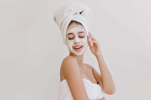 Il ritratto di bella giovane donna che si diverte con gli asciugamani dopo fare il bagno fa la maschera cosmetica sul viso.