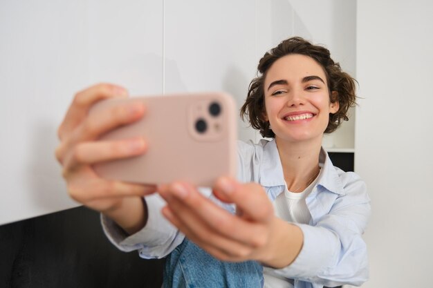 Il ritratto di bella ragazza castana prende il selfie sullo smartphone nella sua cucina che posa per la foto su mo