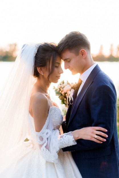 Il ritratto di bella sposa e sposo con gli occhi chiusi sta abbracciando vicino all'acqua all'aperto la sera