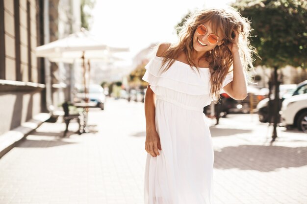 Il ritratto di bello modello biondo sveglio sorridente dell'adolescente senza trucco in vestito bianco dai pantaloni a vita bassa dell'estate copre la posa sui precedenti della via