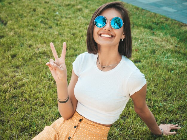 Il ritratto di bello modello castana sorridente si è vestito in vestiti dei pantaloni a vita bassa dell'estate. Ragazza d'avanguardia che si siede sull'erba nel parco. Divertimento positivo e divertente della donna Mostra il segno di pace