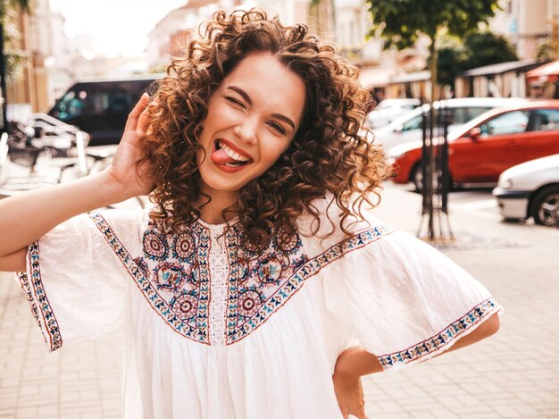 Il ritratto di bello modello sorridente con l'acconciatura dei riccioli di afro si è vestito in vestito bianco dai pantaloni a vita bassa dell'estate.