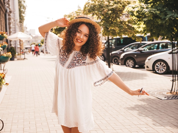 Il ritratto di bello modello sorridente con l'acconciatura dei riccioli di afro si è vestito in vestito bianco dai pantaloni a vita bassa dell'estate.