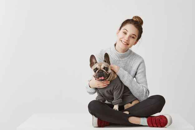 Il ritratto di giovane adulto felice di buon umore che si siede sulla tavola a casa ha attraversato le gambe. Casalinga felice che accarezza bulldog francese con piacere. Copia spazio, concetto di famiglia