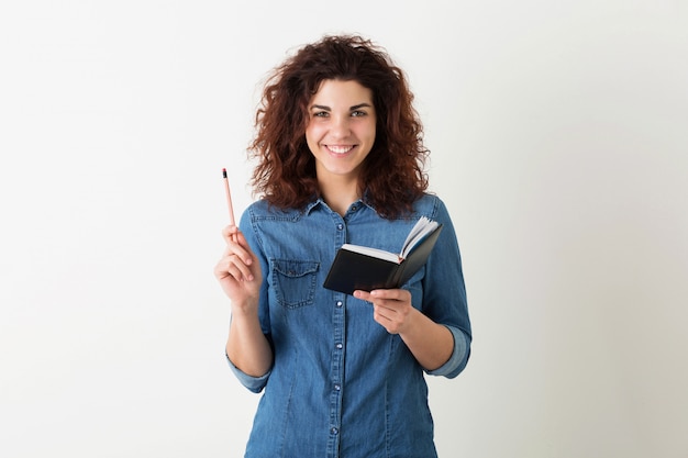 Il ritratto di giovane donna graziosa sorridente naturale con l'acconciatura riccia in camicia del denim che posa con il taccuino e la penna ha isolato, l'apprendimento dello studente, avendo idea