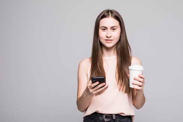 Il ritratto di giovane donna sorridente nel messaggio mandante un sms della camicia sul telefono cellulare e sulla tenuta della tazza di caffè per andare ha isolato sopra la parete grigia