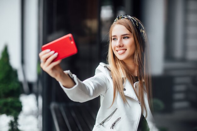 Il ritratto di giovane femmina bionda fa un selfie dal telefono rosso nella via.