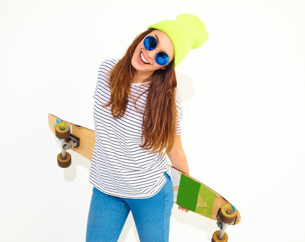 Il ritratto di giovane modello alla moda della donna in estate casuale copre in cappello giallo del beanie che posa con lo scrittorio di longboard. Isolato su bianco