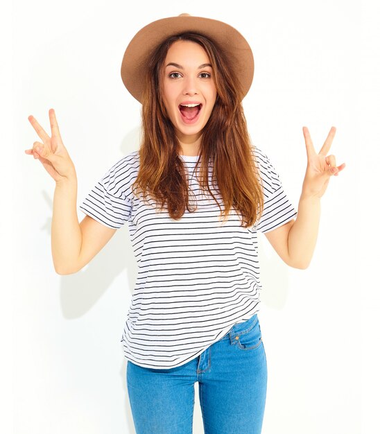 Il ritratto di giovane modello di risata alla moda della donna in estate casuale copre in cappello marrone con trucco naturale isolato sulla parete bianca. Mostra del segno di pace