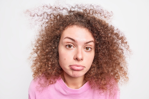 Il ritratto di indignato confuso con il labbro inferiore delle borse dei capelli ricci sembra sorprendentemente imbarazzato mentre sente notizie inaspettate vestito con un maglione casual isolato sul muro bianco