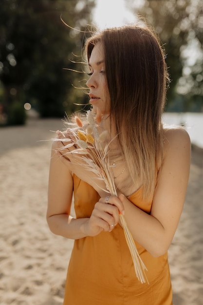 Il ritratto di profilo della bella ragazza europea con l'acconciatura scura sta guardando da parte e tiene in mano l'erba di cotone che cammina sulla spiaggia sabbiosa al sole