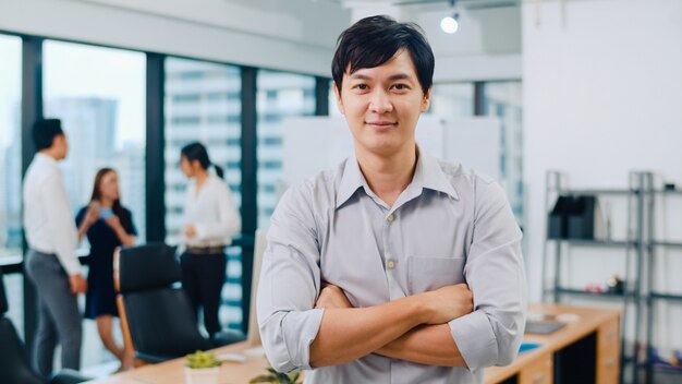 Il ritratto di riuscito abbigliamento casual astuto dell'uomo d'affari esecutivo bello che esamina la macchina fotografica e che sorride, armi ha attraversato nel posto di lavoro moderno dell'ufficio. Giovane ragazzo dell'Asia che sta nella sala riunioni contemporanea.