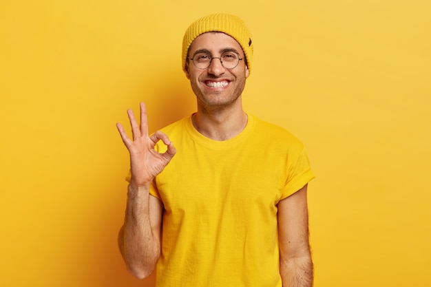 Il ritratto di un bel giovane fa il gesto giusto, dimostra accordo, ama l'idea, sorride felice, indossa occhiali da vista, cappello giallo e maglietta, modelli al coperto. Va bene, grazie. Segno della mano