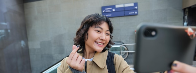 Il ritratto di un'elegante studentessa coreana si fa un selfie per strada, tiene lo smartphone e si mette in posa sul suo