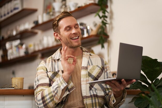 Il ritratto di un giovane collega sorridente che si unisce a una riunione online da una caffetteria mostra le chat video sul portatile