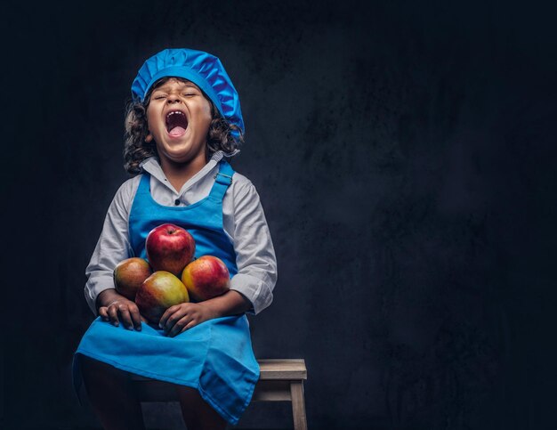 Il ritratto di un ragazzino divertente con capelli ricci marroni vestito con un'uniforme da cuoco blu tiene le mele sedute su uno sgabello di legno in uno studio. Isolato sullo sfondo scuro con texture.